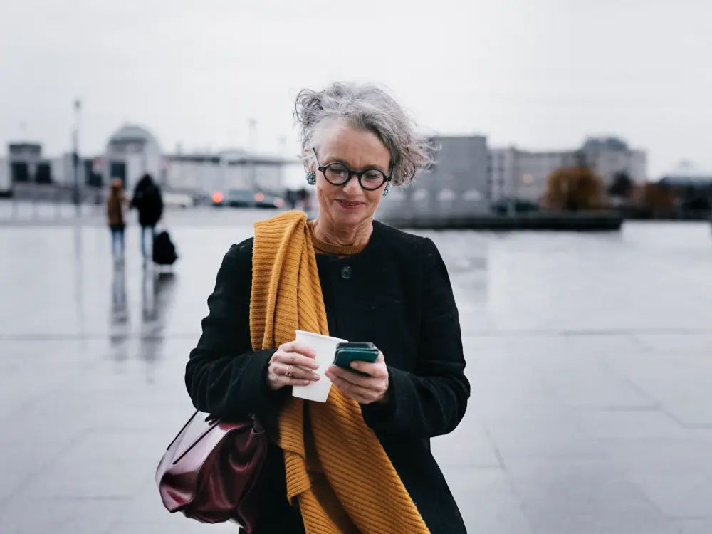 Ältere Frau mit Brille und gelbem Schal steht auf einem Platz und schaut auf ihr Smartphone
