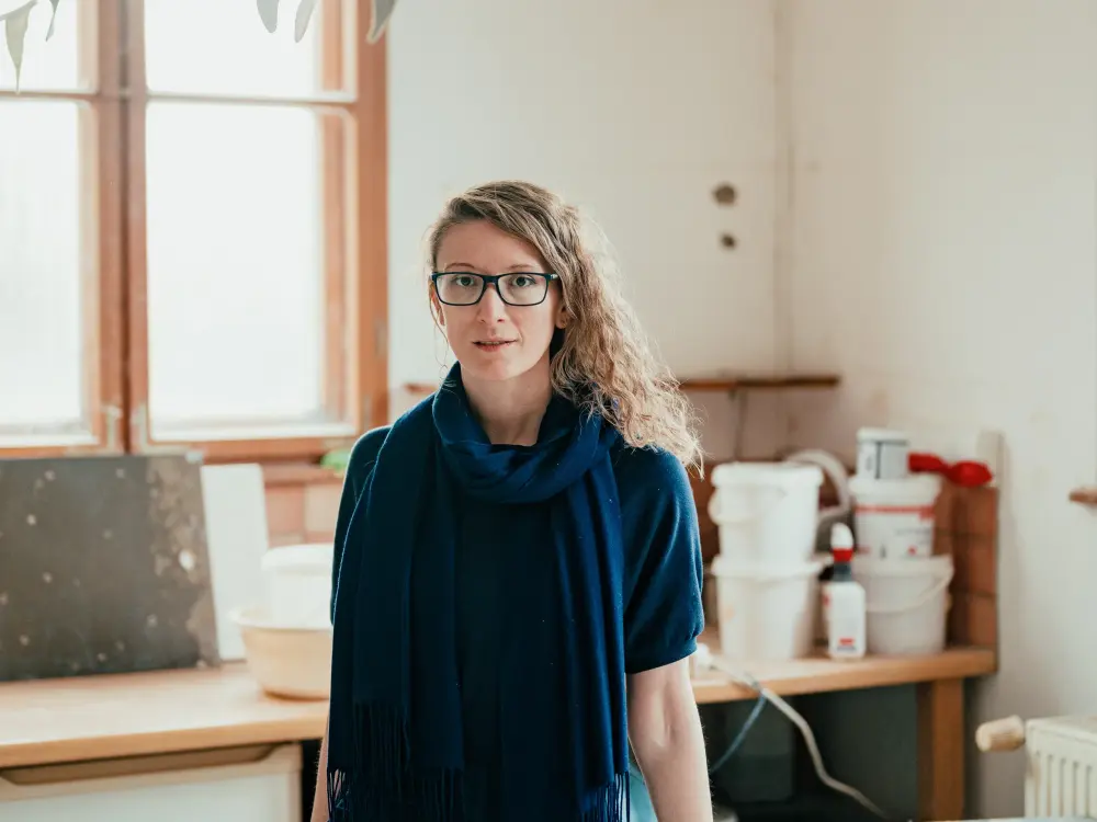 Frau in blauem Kleid und Schal steht an einem Tisch in einem Atelier
