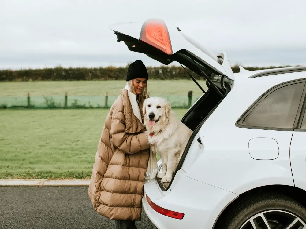 Frau mit goldenem Retriever steht neben einem Auto mit offenem Kofferraum.