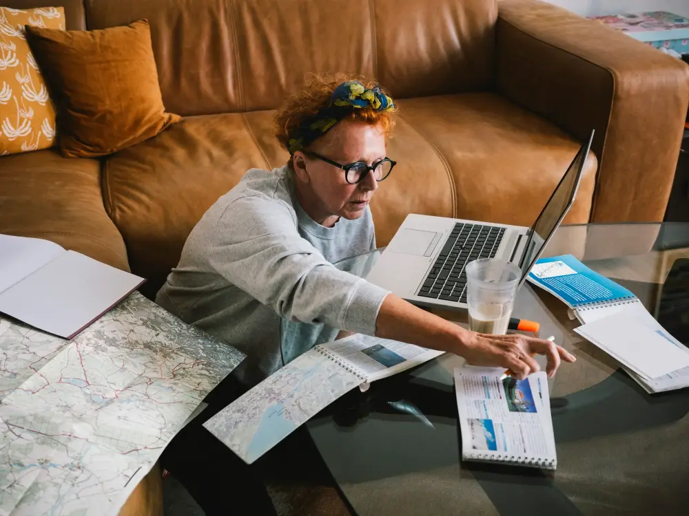 Frau mit Brille plant eine Reise mit Laptop, Karten und Reiseführern.