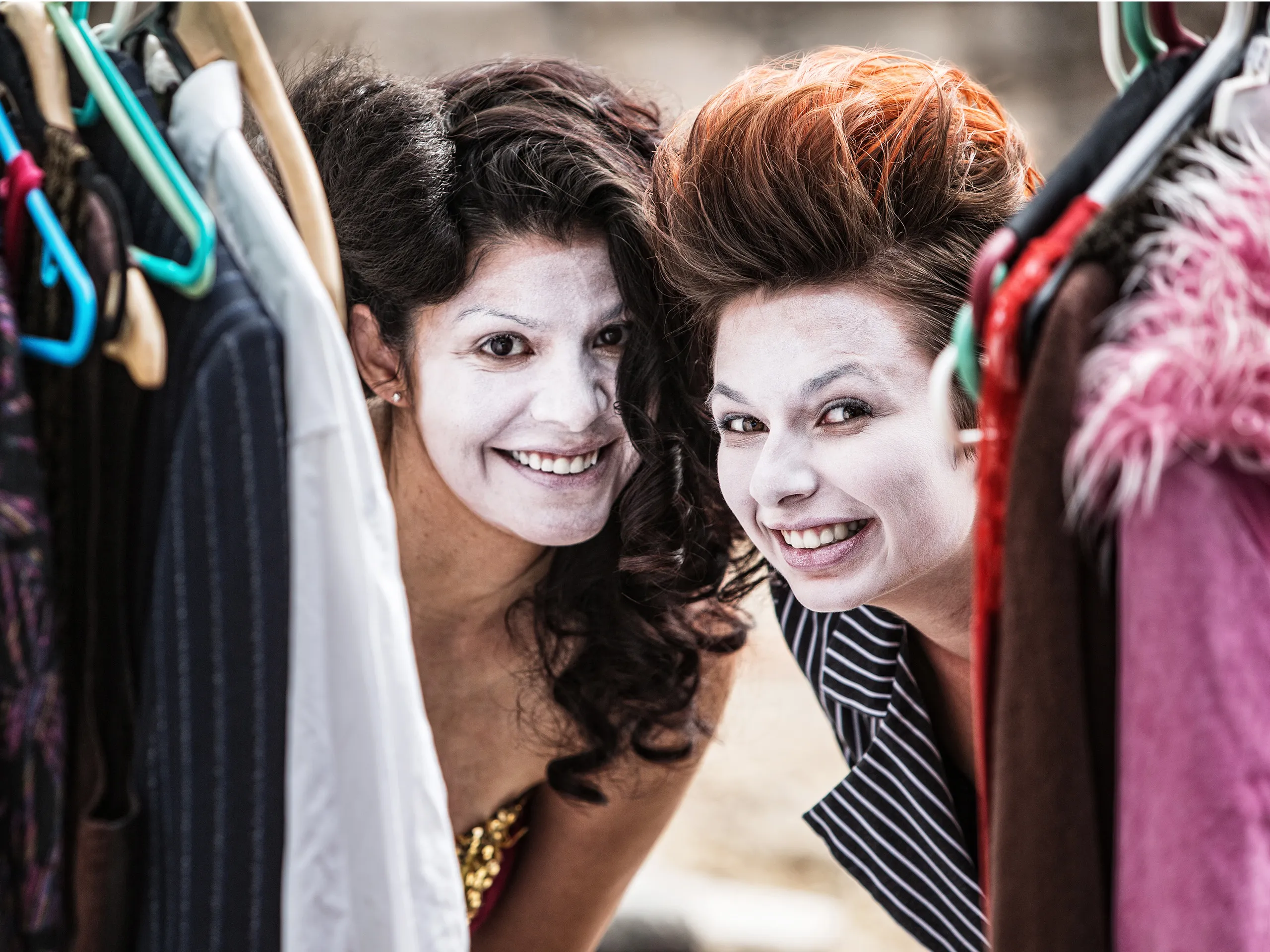 Zwei Frauen in der Garderobe schauen zwischen Kleiderbügeln in die Kamera