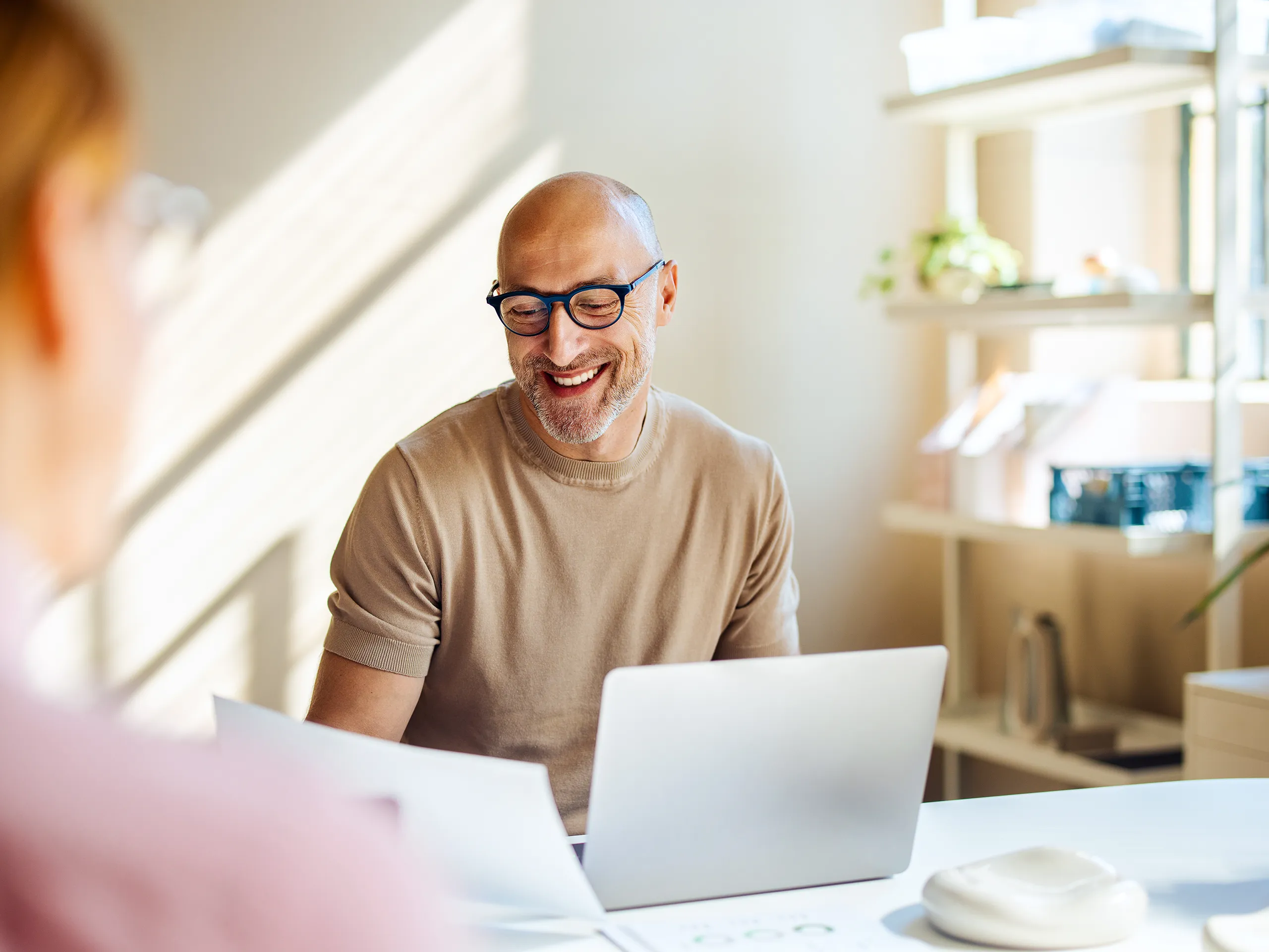 Lachender Mann mit Brille arbeitet mit Laptop und Dokumenten an einem Schreibtisch.