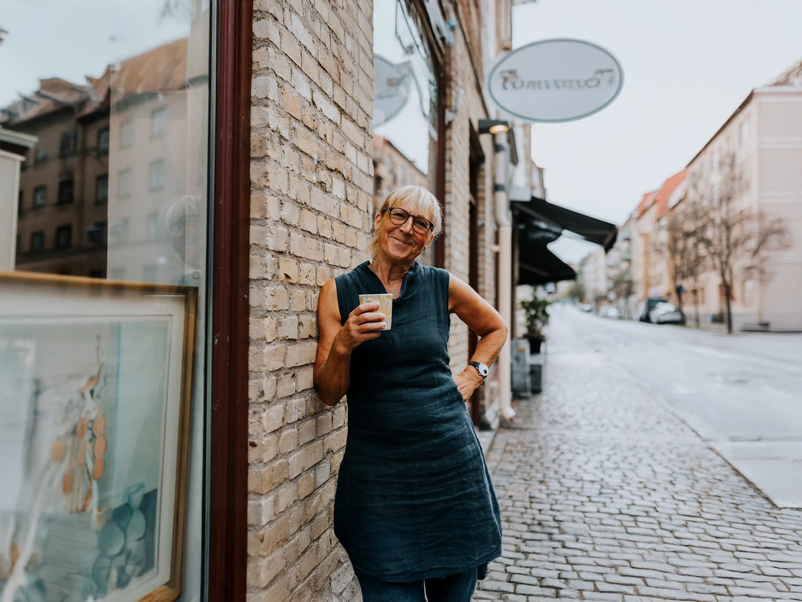 Ältere Frau mit Kaffee lehnt lächelnd an Hauswand auf leerer Straße.