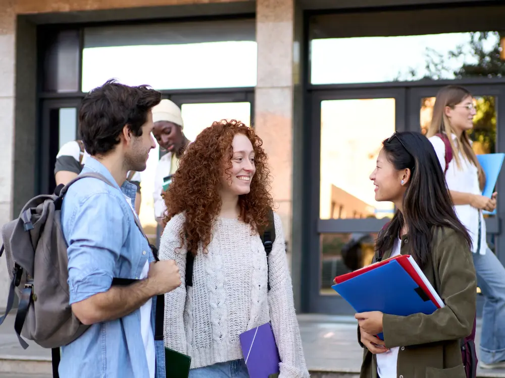 Drei junge Menschen unterhalten sich lächelnd vor einem Campusgebäude.