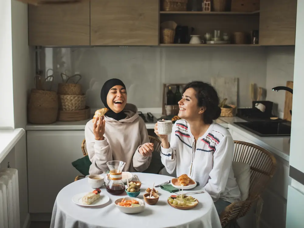 Zwei Frauen frühstücken lachend an einem gedeckten Tisch in der Küche