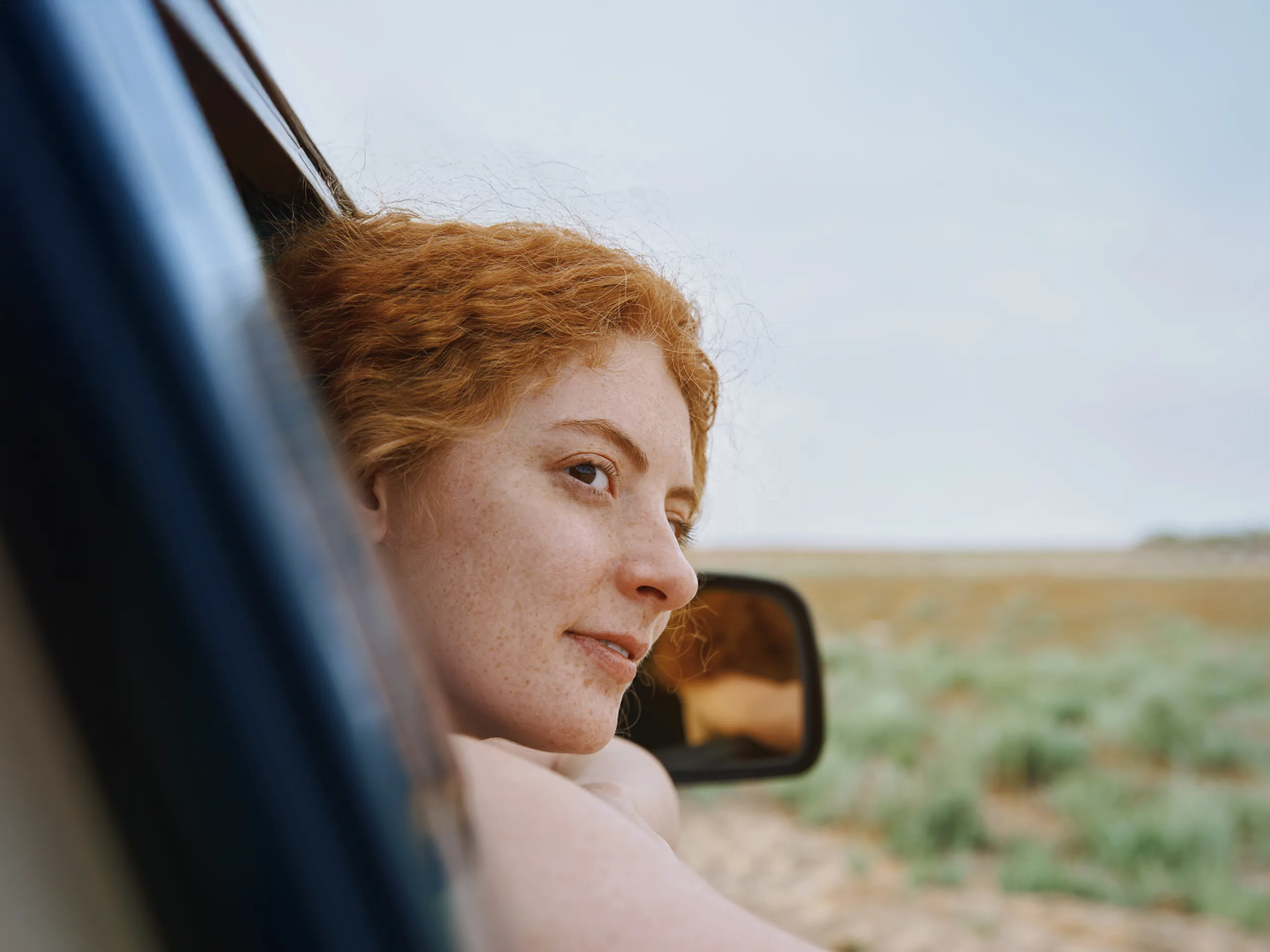 Frau mit roten Haaren lehnt sich aus dem Autofenster und genießt die Landschaft.