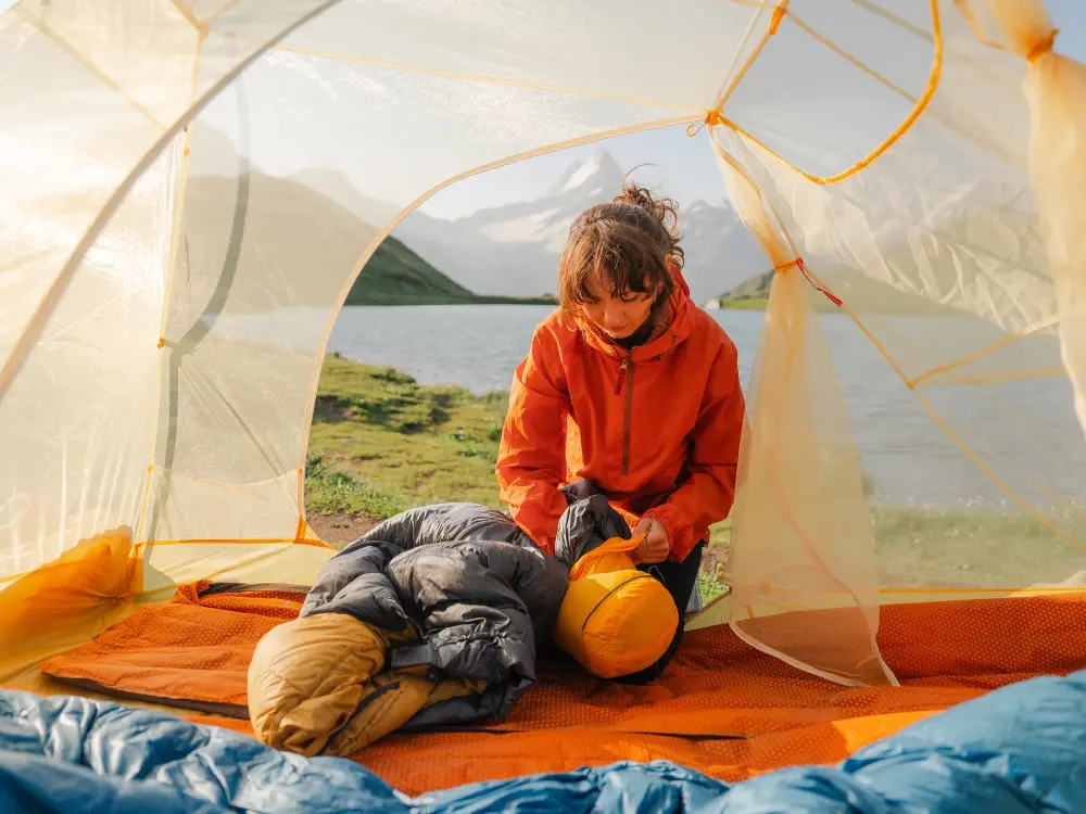 Frau im orangefarbenen Outdoor-Set packt einen Schlafsack in einem Zelt am See ein.